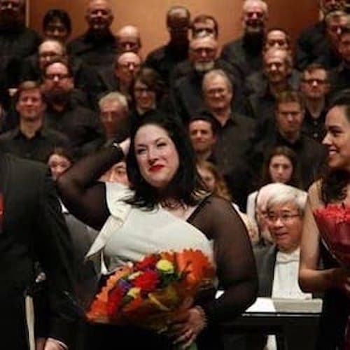 Three people standing on stage in front of a chorus. They are each holding huge bouquet in their arms.