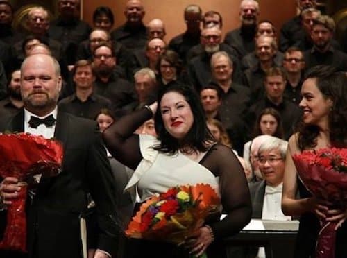 Three people standing in front of a choir on stage. Leah is in the center and all three are holding bouquets of red flowers.